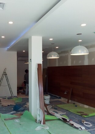 Interior en proceso de remodelación para un centro de ventas del Grupo Centenario, mostrando una pared con revestimiento de madera, cascos de seguridad de obra sobre una mesa y luminarias modernas instaladas.
